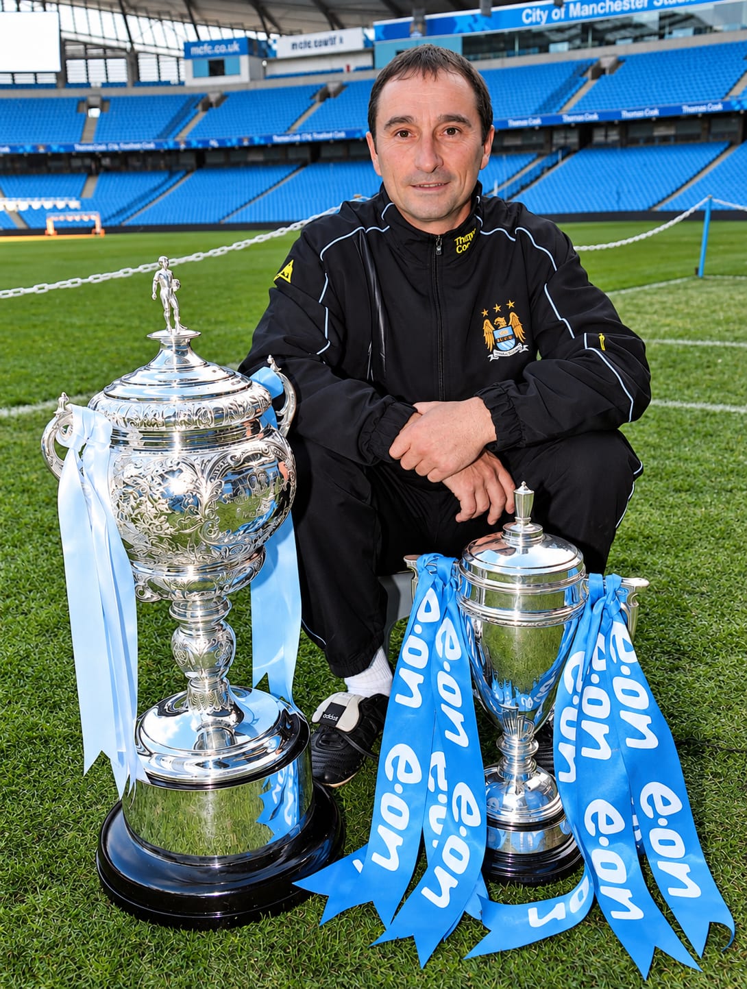 Pete Lowe in een trainingspak van Manchester City, geknield op het veld van het City of Manchester Stadium tussen twee zilveren bekers
